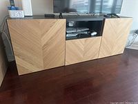 Straight-on view of the IKEA TV unit showing chevron wood design doors and black top surface with a TV on top.