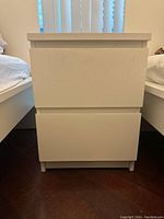 Front view of white side table showing two drawers with no visible handles, standing between two beds.