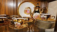 Full set displayed including plates, teacups, saucers, and teapot with clear gold trim and decorative romantic figure detail