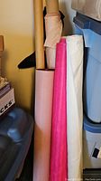 Three bolts of fabric including light pink polyester, bright pink taffeta, and off-white vinyl fabric standing upright between containers.