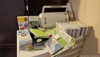 Photo showing Cricut machine from an angled view with cutting mats, various Cricut cartridges, tool cases, and instruction guides on a plastic container.