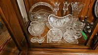 Full assortment of crystal and glassware displayed on a wooden cabinet shelf, including candle holders, creamer, sugar bowl, salt cellars, vase, and decorative plates.
