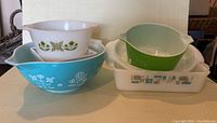 Overview of vintage mixing and serving bowls grouped showing turquoise blue FireKing bowl with white floral design, white and green Anchor Hocking bowls, white Pyrex dish with blue and brown pattern, and green Crate & Barrel berry server bowl.