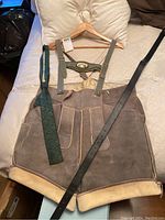 Traditional brown leather lederhosen shorts with cream trim and attached suspenders laid flat on a bed, alongside a green tie and black leather belt.