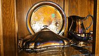 Silverplate lidded dish, pitcher, sauce bowl in front of round ornate serving tray on a wooden shelf