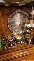 Photo showing silverplate candle holders, creamer, sugar bowl, baby cup, beverage carafe with warming tray, and large tray spaced on a wooden surface, all with tarnished patina.