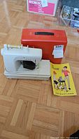 Full view of the white child's sewing machine with spool, red carrying case, and instruction booklet on wooden floor.