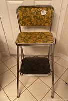 Front view of the folded stool showing the floral patterned padded seat and backrest with a chrome metal frame and black step.