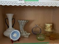 Photo showing all items on wooden shelf including Wedgwood plate, white vase with black handles, metal vase, glassware, candle holder and glass decoration.