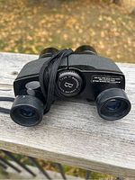 Front view of Bushnell 7x26 binoculars showing lenses, focusing wheel, and neck strap on wooden surface.