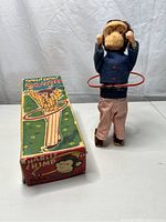 Side view of Charlie The Chimp Hula Expert mechanical toy standing with hula hoop, next to the original colorful illustrated box showing the character in a dancing pose with a hula hoop.