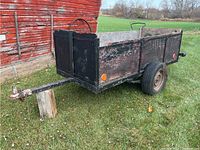 Full side and front angle view of homemade utility yard trailer with visible tongue and tires on grass near red barn.