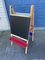 Front view of the wooden kids art easel showing the black chalkboard side with a red tray attached at the bottom for supplies.