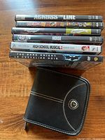 Six DVD movies stacked on a wooden surface with their original cases, plus a black zippered disc holder visible in front.