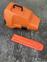 Orange hard plastic Stihl chainsaw case and bar cover shown outdoors on concrete, case with black handle and latches.