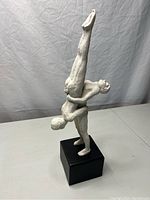 Side view of two nude male gymnasts sculpture on black base, showing muscular figure holding inverted legs of second figure.