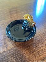 Image showing black porcelain trinket dish with a small brown fish figurine jumping out from the edge on wooden surface.