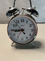 Front view of a metal wind up alarm clock with white face, black numerals, and brass colored hands including a red second hand with a green tip alarm hand.