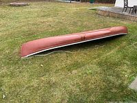 Full side view of the red fibreglass canoe resting upside down on grass, showing the length and white trim along the bottom edge.