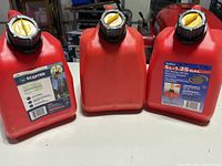 Three red 5-litre self-venting gasoline containers with yellow and black caps, front view showing labels on two containers.