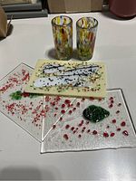 Five handmade art glass items arranged on white surface: three glass panels in front and two shot glasses behind, showing color and pattern details.