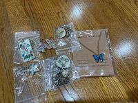 Photo showing 5 necklaces with different pendants in plastic bags including a blue butterfly necklace from Make A Wish and a pair of aura finish angel earrings.