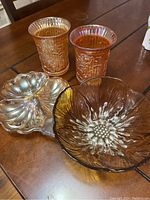 Photo displaying two iridescent Carnival Glass vases/glasses, amber sunflower-shaped bowl, and three-piece matching glass set including creamer, sugar bowl, and small dish.