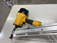 Photo of yellow and black Powerfist air-powered framing nailer with metal magazine and red nail guide tip laid on a white table surface.