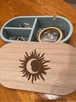 Open blue Bizou jewelry box showing three compartments filled with various costume jewelry including rings, bracelets, earrings, brooches, and necklaces. The wooden lid features a sun and moon design.