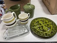 Overview of all items in the lot placed on a white table with boxes in background: glass butter dish, green cast iron trivet, green sugar dish with lid, cream/milk pitcher, and striped sugar bowl and pitcher.