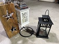 Three decorative lantern items arranged on floor: black metal votive lantern, white metal lantern with intricate design and battery candle inside, wooden panel with star cutouts and black battery-operated light strand