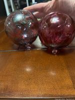 Pair of hand blown glass balls featuring bubble textured surfaces and pink/purple iridescent colors, sitting on a wooden surface.