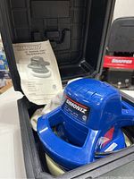 Blue Simoniz random orbit polisher with dual handles placed inside black hard carrying case alongside instruction manual and polishing pads.