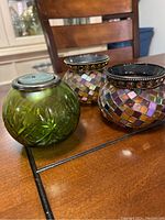 Photo of two mosaic glass tealight holders with multicolored square pieces and one green cut glass holder with metal lid placed on a wooden table.