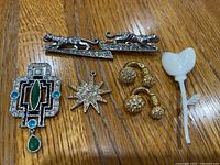 Five vintage jewelry pieces placed on wooden surface including two silver-tone running panther brooches with rhinestone details, one geometric art deco style brooch with colored stones, one gold-tone starburst pendant with rhinestones, and one white opaque floral pendant.