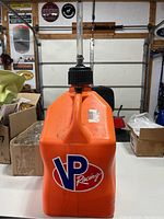 Front view of orange VP Racing 5 gallon fuel jug with large black screw-on cap and clear flexible filling spout attached.