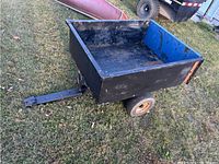 Side and top angle view of black metal yard trailer with blue interior panels on grass. Shows general shape and condition.
