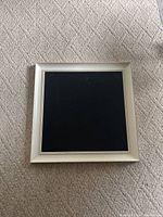 Photo of a square black chalkboard with white frame lying on carpeted floor.