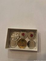 Top-down photo of six commemorative coins in a white box. Coins include silver-tone and one gold-tone coin, some with red poppy emblems and Queen Elizabeth II portraits.