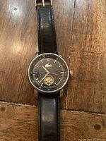 Front view of Lacoste automatic watch with black dial and visible balance wheel on wooden surface.