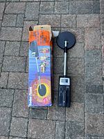 Metal detector next to its original box on a brick surface.