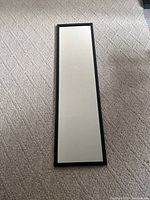 Rectangular wall mirror with black frame lying on beige/carpeted floor. Mirror surface is clear and frame is in good condition.