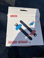 Box front showing product image of two black armband hand straps with blue and red Joy-Con controllers attached, brand Aolion, text 'HAND STRAP x2'