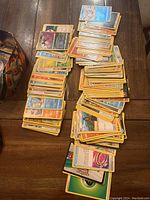 Multiple stacks of assorted Pokémon cards with visible character cards and energy cards on wooden surface, next to a Pokémon-themed tin container.