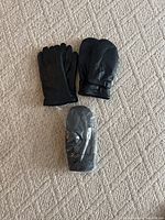 Three pairs of black leather handwear including two pairs of gloves and one pair of mittens, one still in packaging, on carpeted surface.
