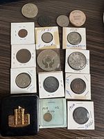 Top view of multiple coins, some in holders with dates and values annotated, including a wooden nickel token and a metal castle figurine in box.