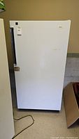 White upright freezer closed, showing overall exterior with minor wear and a vertical handle on the left.