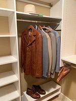 Brown leather jacket and 4 men's suits hanging on closet rod with brown Hush Puppies shoes and straw hat on shelves