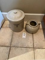 Two beige ceramic pottery pieces on tiled floor: a lidded Alberta Potteries No 2 crock and a Medalta handled pot.