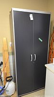 Closed metal storage cabinet with black doors, silver trim, and white handles. Cabinet is tall and slim, placed in a garage with some items nearby.